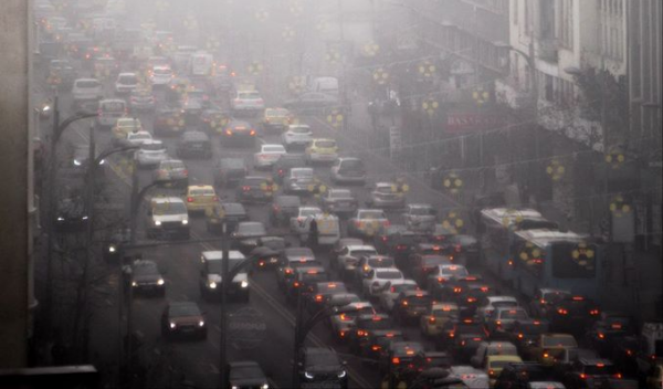 Hundreds of vehicles sit in heavy traffic within a bustling city, while surrounded by heavily polluted air